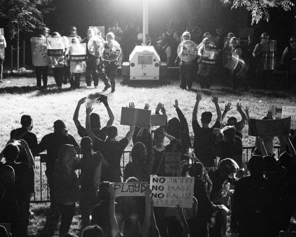 A black and white photo of Black Lives Matters protestors from behind with their hands up as police officers stare them down in riot gear